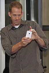 La imagen de un hombre sosteniendo un teléfono blanco. El usa una camisa café a rayas.