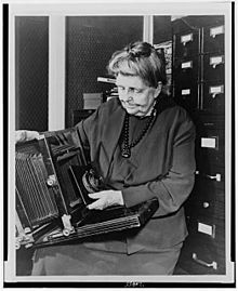 Frances Benjamin Johnston, three-quarter length portrait, holding and looking down at camera, facing slightly left LOC 5890981070.jpg