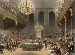 View of a large room, showing the Speaker of the House sitting at the end. Down each side of the room, MPs are sitting – one MP is standing on the right, giving a speech. Balconies are on either side, with spectators visible.