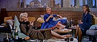 Monroe en How to Marry a Millionaire. Lleva un traje de baño naranja y está sentada junto a Betty Grable, que lleva pantalones cortos y una camisa, y Lauren Bacall, que lleva un vestido azul.