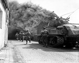 United States Army soldiers supported by a M4 Sherman tank move through a smoke filled street in Wernberg, Germany during April 1945