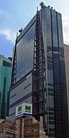 Ground-level view of a blue, glass, rectangular high-rise; attached to one side of the building are two structures consisting of poles that run the length of the building