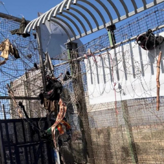 Remnants of clothing, wooden sticks and backpacks stuck to the barbed wire over the doors and on the fence (cropped).png