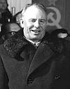 A man wearing a long coat, smiling, in front of a group of people and the Soviet flag