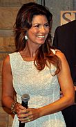 Shania Twain con un vestido blanco está de pie sosteniendo un micrófono y sonriendo.